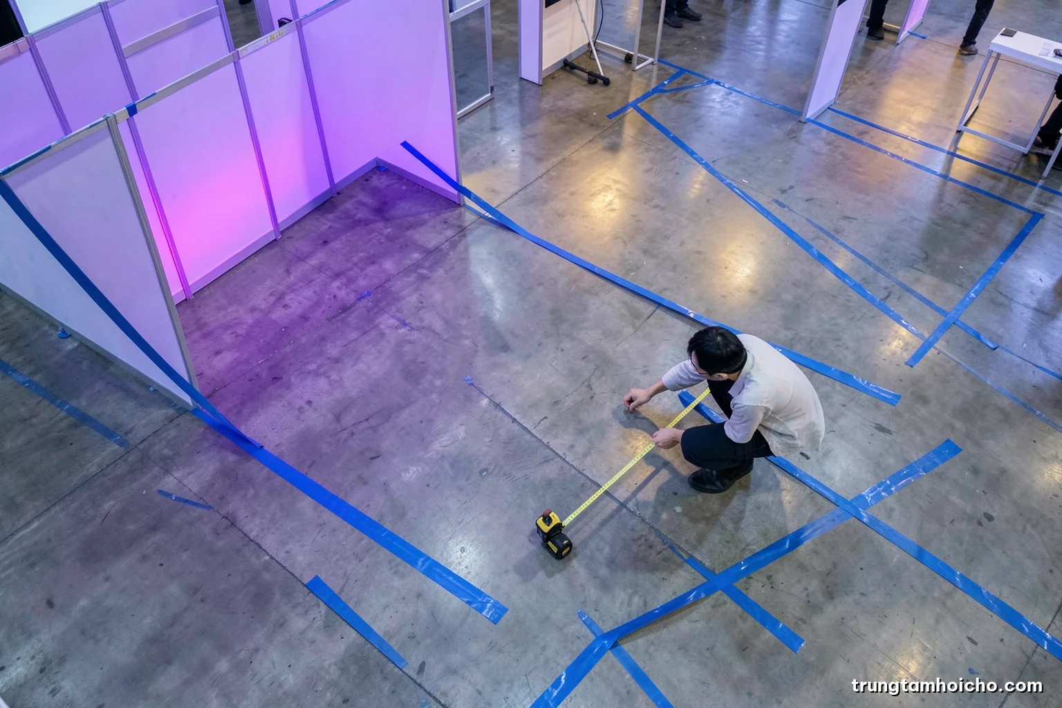 Overhead view of blue masking tape outlining booth zones on a polished concrete floor, a Vietnamese designer kneeling with a tape measure, soft diffused light, blue and concrete grey palette