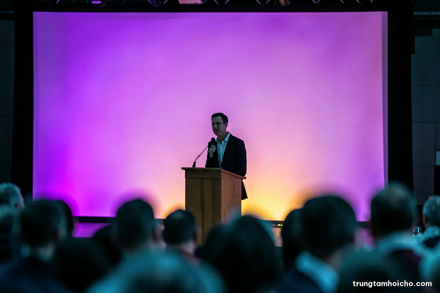 Silhouette of a speaker standing at a wooden lectern against a large blank projection wall, audience heads softly blurred in foreground, cool teal and charcoal palette, editorial photography