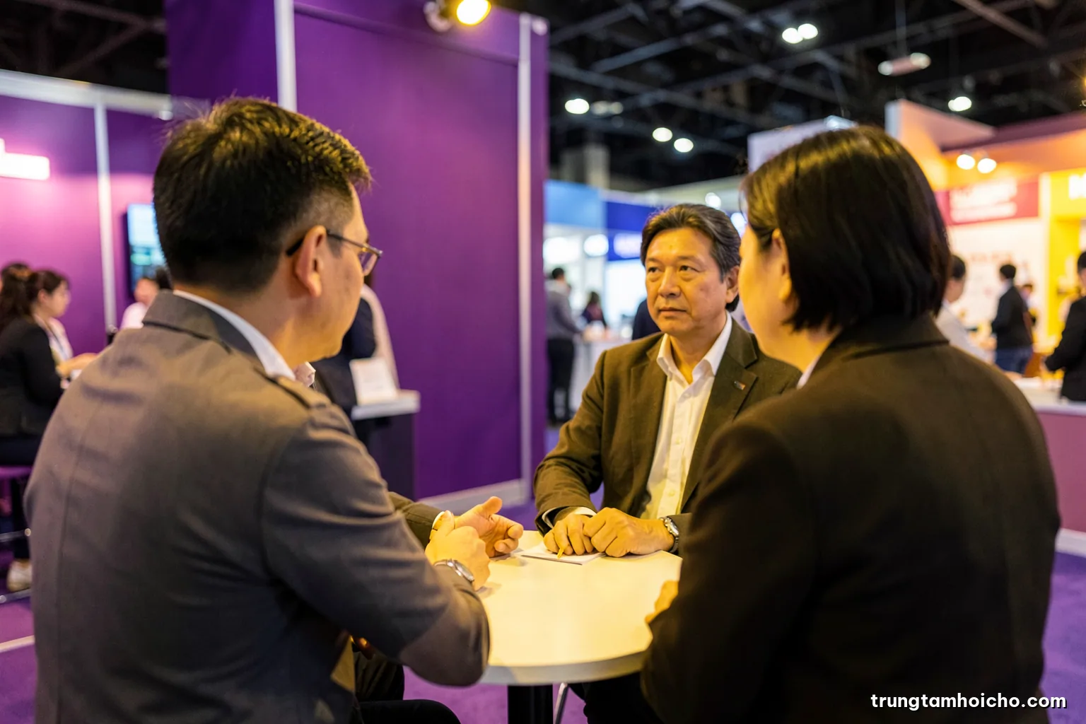 Over-the-shoulder view of a Vietnamese engineer listening intently to a visitor across a small round table inside a trade fair booth, warm tungsten light, olive and plum palette