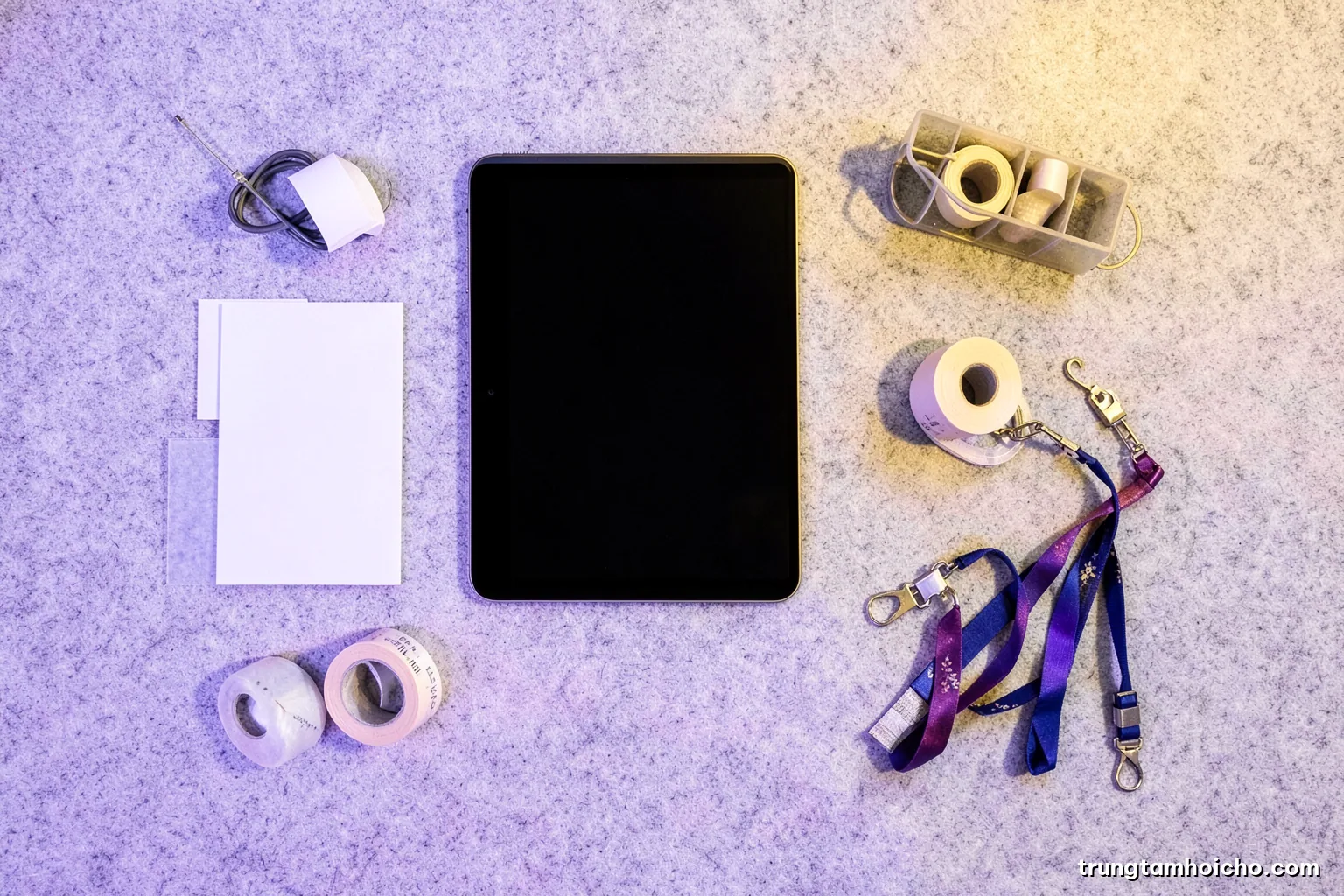 Top-down flat lay of neatly arranged exhibition essentials on light grey felt: blank business cards, a tablet with dark screen, cable organizer, tape rolls, lanyards, soft daylight, minimal palette