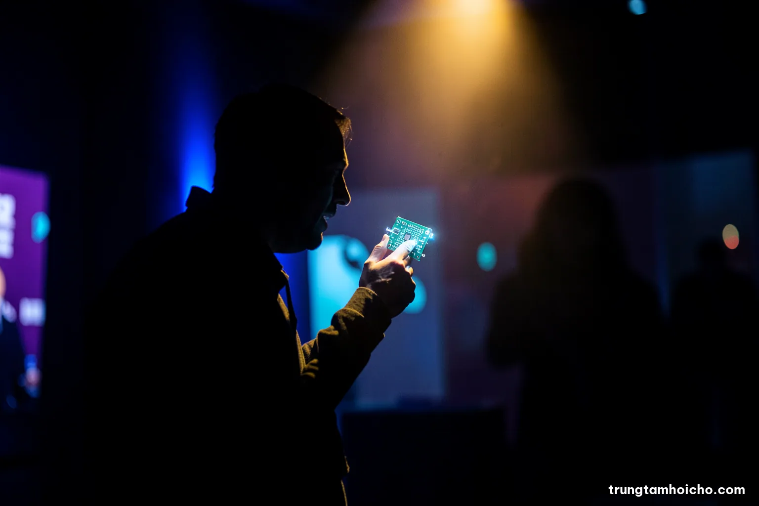 Silhouette of a visitor examining a small circuit board under a spotlight in a dark exhibition booth, blue rim light and soft bokeh, deep navy and electric cyan palette, technology editorial photography