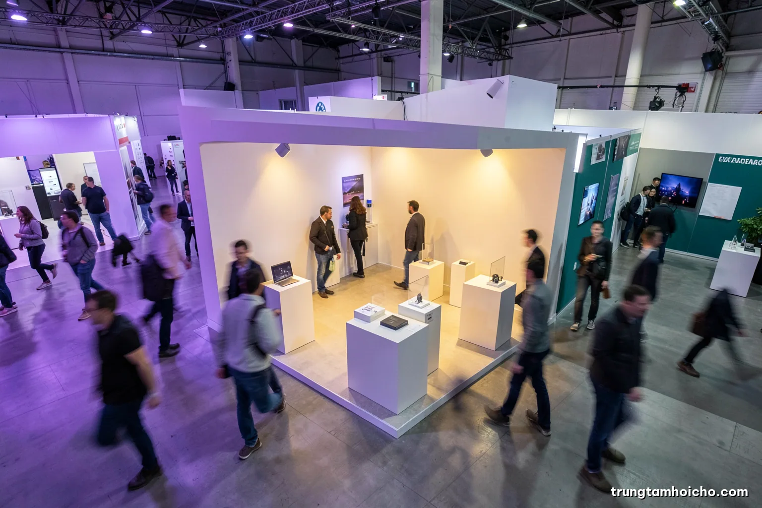 High angle view of an exhibition aisle where visitors slow down at a minimalist open booth with product plinths, motion blur on passersby, neutral grey and teal palette