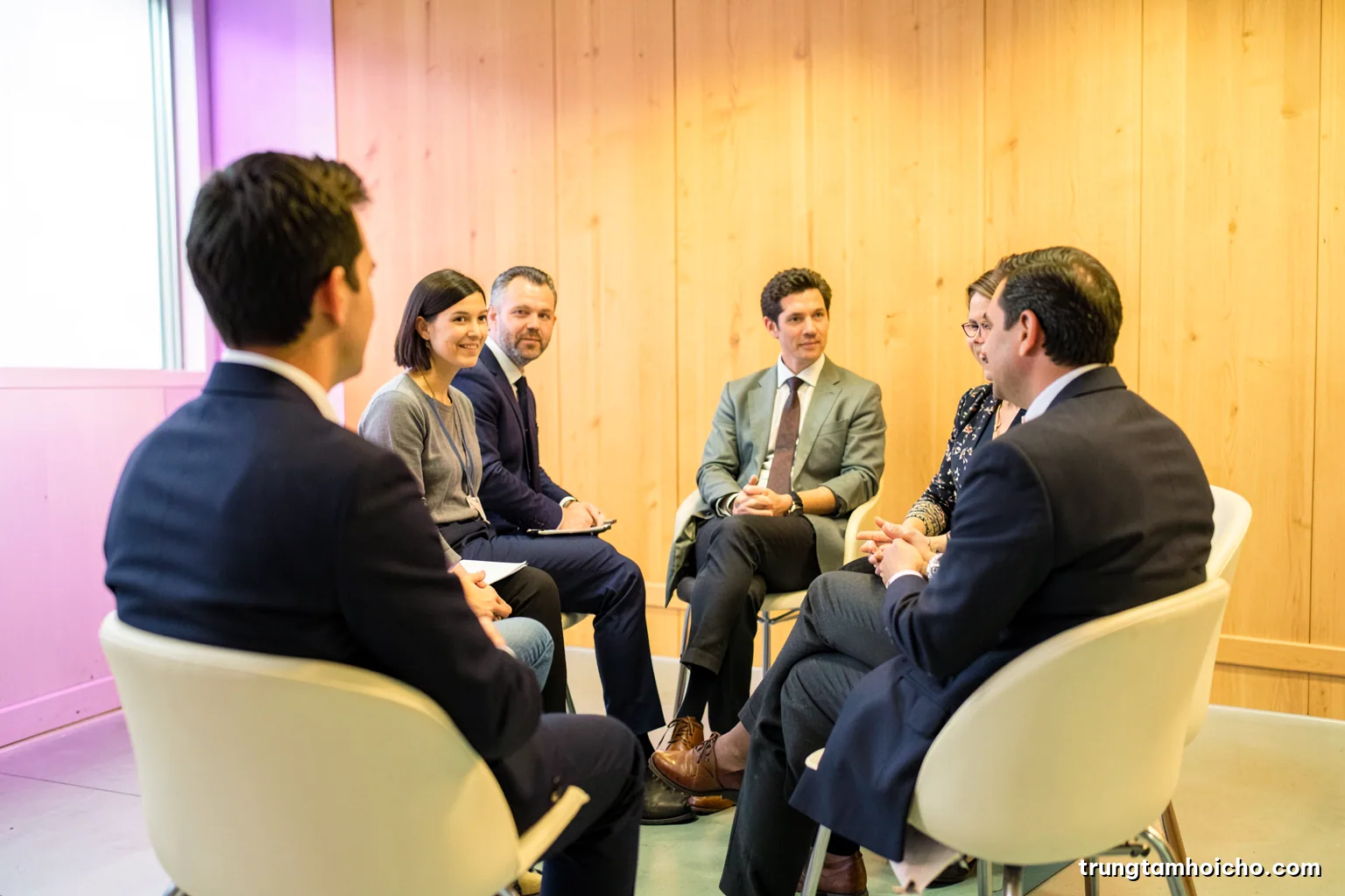 Over-the-shoulder view of a small circle of chairs in a bright wood-panelled room, five participants in quiet discussion, soft daylight, sage green and cream palette, editorial photography