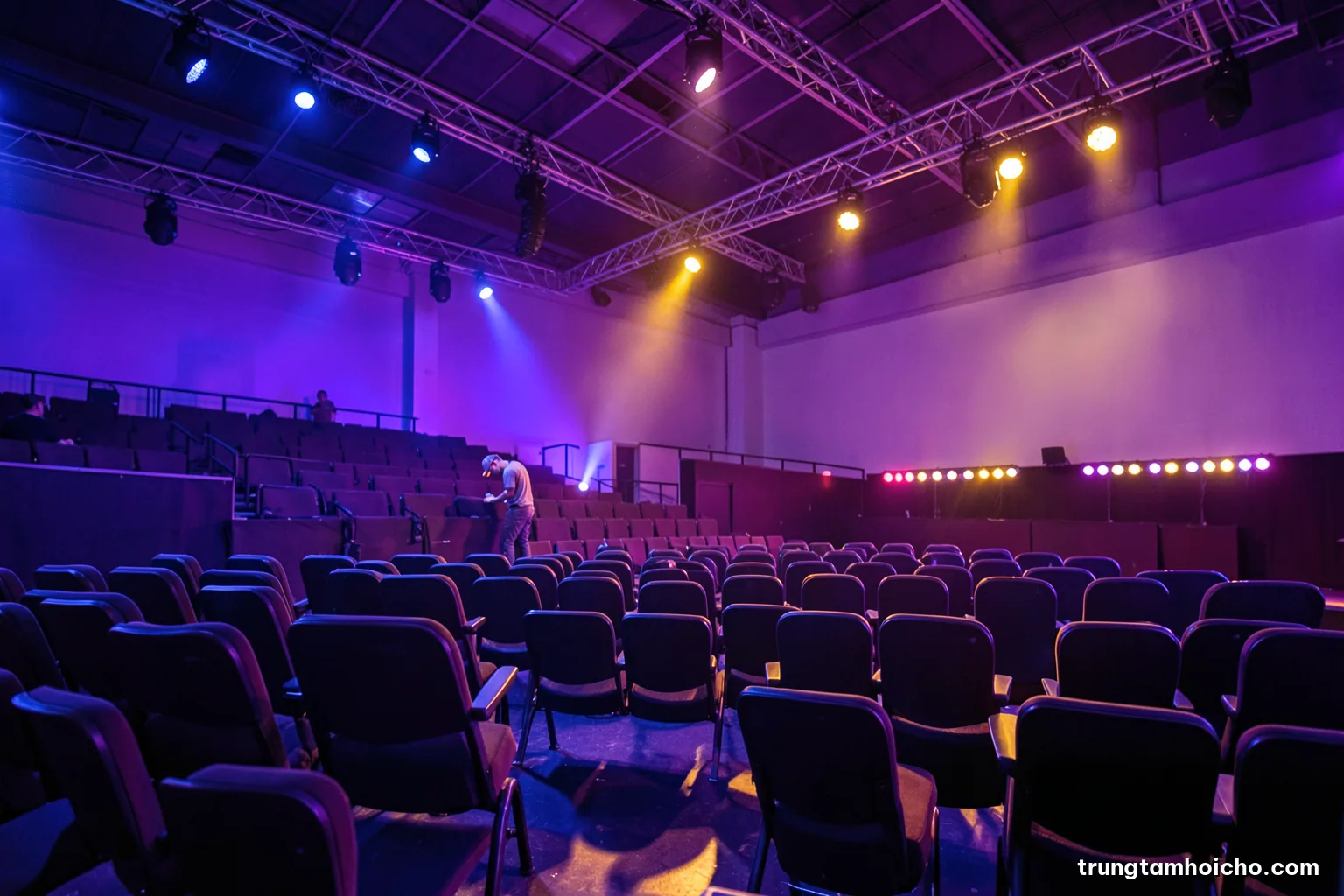 Low angle view of an empty conference auditorium before an event, rows of chairs, a single technician adjusting stage lights, cool blue and amber lighting