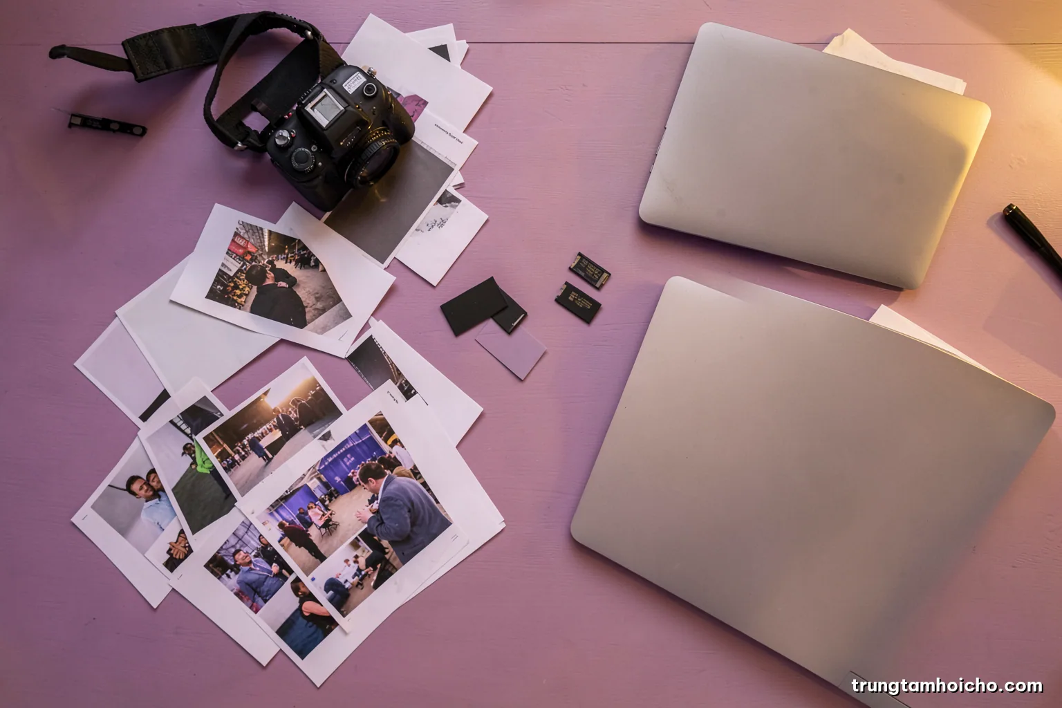 Top-down view of an editing desk with scattered unlabeled photo prints from a trade fair, a camera, memory cards and a closed laptop, soft overcast light, warm neutral palette