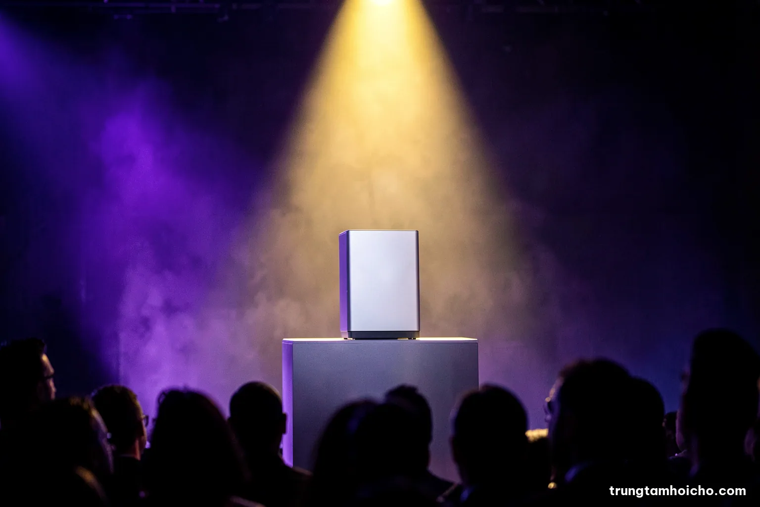 Low-angle shot of a sleek unbranded product on a pedestal under a single spotlight, dark stage with faint haze, silhouettes of audience heads in foreground, cobalt and silver palette