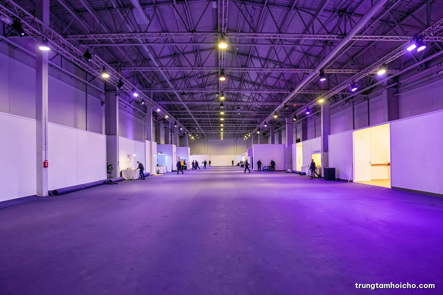 Wide shot of an empty exhibition hall after the fair closes, a few staff rolling up carpet under high industrial lights, long perspective, cool grey and violet tones
