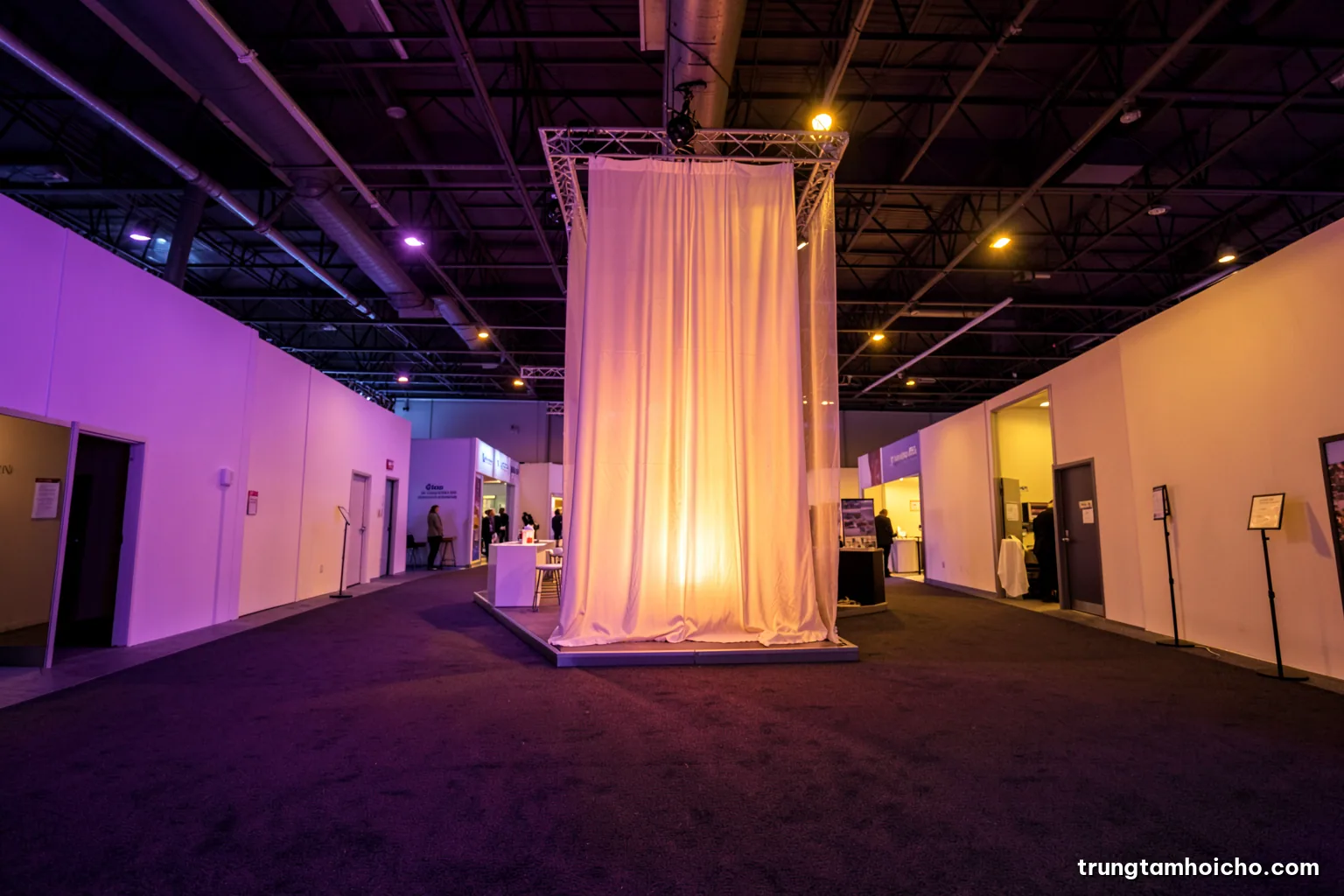 Low angle view from an empty exhibition aisle toward an island booth with a tall fabric tower glowing softly, carpet in the foreground, dusk-like warm hall lighting, amber and charcoal palette