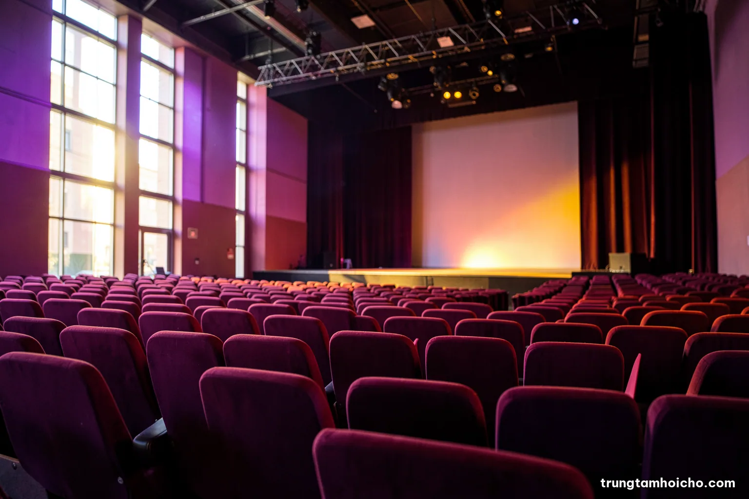 Low angle view of an empty auditorium with rows of upholstered seats and a softly lit stage, morning sun through tall windows, burgundy and gold palette, editorial photography